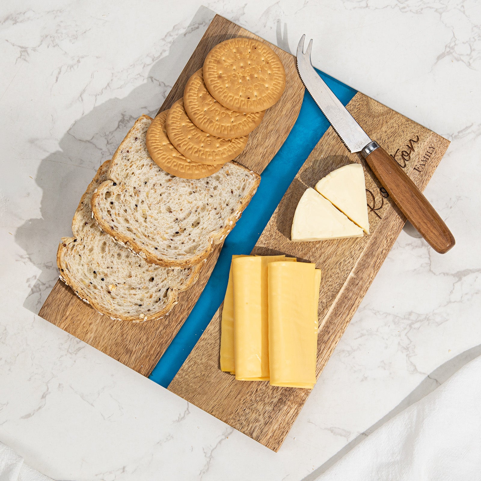 Overhead view of personalized epoxy resin cutting board with cheese bread and crackers charcuterie spread on marble surface blue river acacia wood